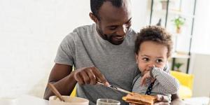 Father with young son on his lap spreading peanut butter on toast.